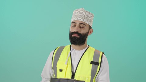 كتابة وتدوبن الملاحظات على ورقة، مفهوم الهندسة والانشاءات المعمارية، بورتريه لمهندس عربي خليجي عماني يرتدي الدشداشة و الكمه وسترة الحماية قاعدة كتابة ينظر إليه بايماءات التركيز والتمعن، مفهوم إدارة المشاريع وتدقيقها، العمل في القطاع الهندسي، خلفية زرقاء