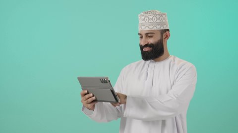متابعة الأفلام والمسلسلات الممتعة، استخدام التكنولوجيا و التقنيات الحديثة, المحادثة مع الأهل والأصدقاء، متابعه أخر أخبار مواقع التواصل، تصفح مواقع التواصل الاجتماعي، بورتريه مقرب لرجل عربي خليجي عماني يرتدي الدشداشة و الكمة يحمل بيده جهاز التابلت، خلفية زرقاء