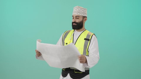 مفهوم الهندسة والانشاءات المعمارية، بورتريه لمهندس عربي خليجي عماني يرتدي الدشداشة و الكمه وسترة الحماية يمسك المخطط ينظر إليه بايماءات التركيز والتمعن، مهنة ووظيفة للشباب في سلطنة عمان، مفهوم إدارة المشاريع وتدقيقها، العمل في القطاع الهندسي، خلفية زرقاء