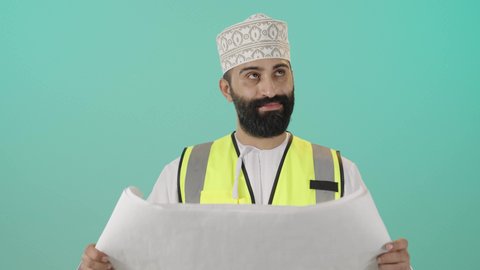 مهنة ووظيفة للشباب في سلطنة عمان، مفهوم الهندسة والانشاءات المعمارية، بورتريه لمهندس عربي خليجي عماني يرتدي الدشداشة و الكمه وسترة الحماية يمسك المخطط ينظر إليه بايماءات التركيز والتمعن، مفهوم إدارة المشاريع وتدقيقها، العمل في القطاع الهندسي، خلفية زرقاء