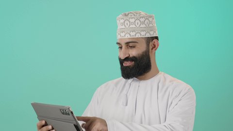 Using technology and modern techniques, chatting with family and friends, following the latest news on social media, browsing social media sites, watching enjoyable movies and series, a close-up portrait of an Arab Gulf Omani man wearing a dishdasha and a kumma holding a tablet in his hand, blue background.