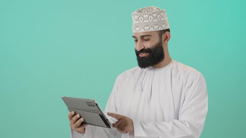 المحادثة مع الأهل والأصدقاء، متابعه أخر أخبار مواقع التواصل، استخدام التكنولوجيا و التقنيات الحديثة, تصفح مواقع التواصل الاجتماعي، متابعة الأفلام والمسلسلات الممتعة، بورتريه مقرب لرجل عربي خليجي عماني يرتدي الدشداشة و الكمة يحمل بيده جهاز التابلت،  خلفية زرقاء