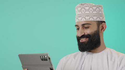 Following the latest news on social media, chatting with family and friends, using technology and modern techniques, browsing social media sites, watching enjoyable movies and series, a close-up portrait of an Arab Gulf Omani man wearing a dishdasha and a kumma holding a tablet in his hand, with a blue background.