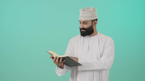 حب المطالعة والقراءة، تنمية الذات خلال أوقات الفراغ بالاطلاع والتعلم، تمضية أوقات الفراغ بالتعلم والتثقيف،الاهتمام بتصفح الكتب المفيدة، بورتريه مقرب لرجل عربي خليجي عماني يرتدي الدشداشه و الكمة يحمل بيده كتاب، مفهوم التأمل والتفكير، خلفية زرقاء