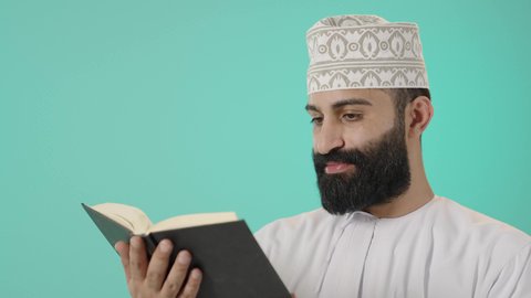 تنمية الذات خلال أوقات الفراغ بالاطلاع والتعلم، حب المطالعة والقراءة، تمضية أوقات الفراغ بالتعلم والتثقيف،الاهتمام بتصفح الكتب المفيدة، بورتريه مقرب لرجل عربي خليجي عماني يرتدي الدشداشه و الكمة يحمل بيده كتاب، مفهوم التأمل والتفكير، خلفية زرقاء