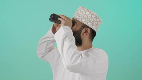المراقبة وانتظار حصول شيء ما، استخدام الأدوات المتطورة لتقريب وتوضيح الرؤية، رؤية الأشياء البعيدة، التخطيط المستقبلي للأهداف والطموحات، حب الاستكشاف والمغامرة، بورتريه لرجل عربي خليجي عماني يرتدي الدشداشة والكمة يمسك منظار ثنائي العينين ، التفكير والتطلع إلى المستقبل, خلفية زرقاء