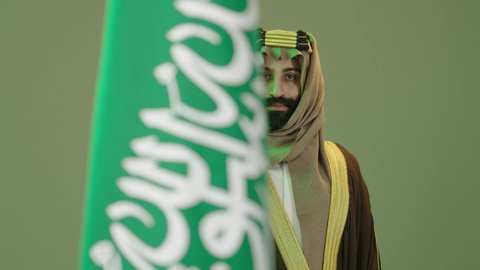 Portrait of a Saudi Arabian Gulf man wearing a bisht, ghutra and agal on Founding Day, holding a sword and standing next to the Saudi flag, the day we began 1727 AD, the anniversary of the founding of the first Saudi state on February 22, Flag Day on March 11, Saudi National Day commemoration on September 23