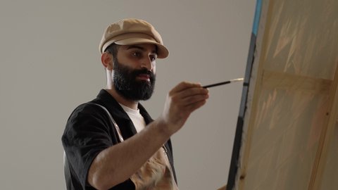 Art workshop, using drawing and painting tools and equipment, mastering the hobby of handicrafts, portrait of a Saudi Arabian Gulf artist wearing traditional clothing and a painting apron sitting on a chair in front of the painting, gestures of joy and happiness