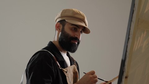 إتقان هواية الفنون اليدوية , استخدام أدوات و معدات الرسم و التلوين , ورشة عمل فنية , بورتريه لرسام عربي خليجي سعودي يرتدي الثوب التقليدي و مريلة الرسم يجلس على الكرسي أمام اللوحة , إيماءات الفرح و السرور