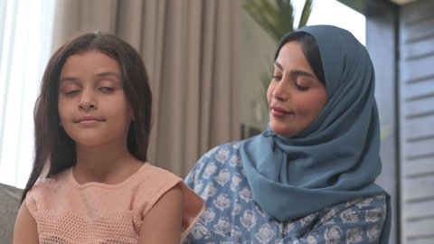 المحافظة على شعر صحي وأنيق، امراة عربية خليجية سعودية ترتدي العباءة والحجاب تجلس على الكنبة تقوم بتصفيف وتمشيط شعر ابنتها باستخدام الفرشاة ( المشط ) ، مفهوم العناية والاهتمام بالأناقة والمظهر الخارجي، إيماءات السعادة والسرور