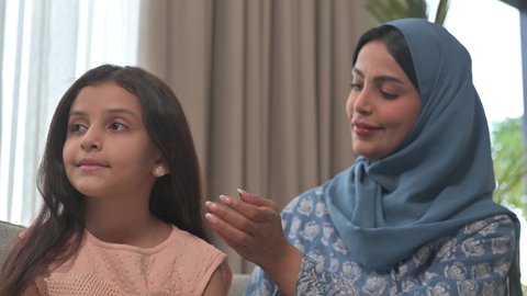 مفهوم العناية والاهتمام بالأناقة والمظهر الخارجي، إيماءات السعادة والسرور، المحافظة على شعر صحي وأنيق، امراة عربية خليجية سعودية ترتدي العباءة والحجاب تجلس على الكنبة تقوم بتصفيف وتمشيط شعر ابنتها باستخدام الفرشاة ( المشط ) 