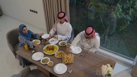 أجواء دينية رمضانية، عائلة عربية خليجية سعودية تجلس حول مائدة الطعام في المنزل، سفرة مليئة بالمأكولات الشهية، اجتماع الأسرة حول مائدة الطعام، افطار الصائم وقت أذان المغرب في رمضان