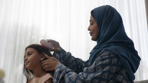  العناية والاهتمام بالأناقة والمظهر الخارجي، إيماءات السعادة والسرور، المحافظة على شعر صحي وأنيق، امراة عربية خليجية سعودية ترتدي العباءة والحجاب تجلس على الكنبة تقوم بتصفيف وتمشيط شعر ابنتها باستخدام الفرشاة ( المشط ) 