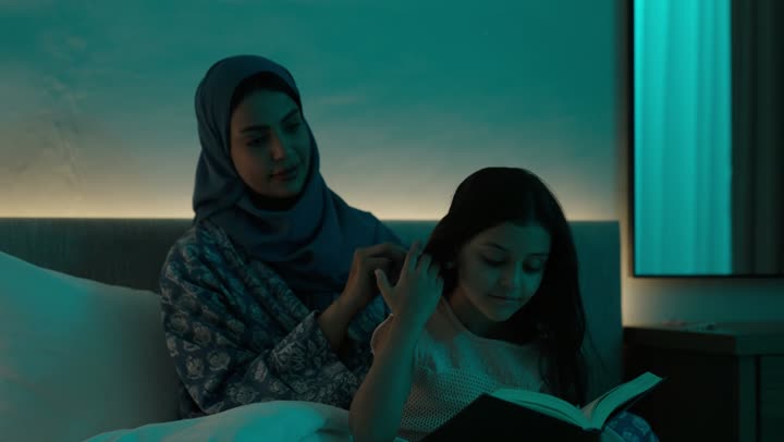 الاهتمام والعناية بالأناقة والمظهر الخارجي، إيماءات السعادة والسرور، المحافظة على شعر صحي وأنيق، امراة عربية خليجية سعودية ترتدي العباءة والحجاب تجلس على السرير تقوم بتصفيف وتمشيط شعر ابنتها باستخدام الفرشاة ( المشط )