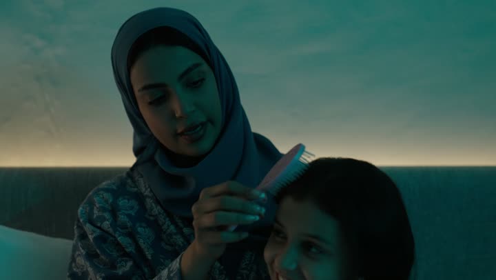الاهتمام والعناية بالأناقة والمظهر الخارجي، إيماءات السعادة والسرور، المحافظة على شعر صحي وأنيق، امراة عربية خليجية سعودية ترتدي العباءة والحجاب تجلس على السرير تقوم بتصفيف وتمشيط شعر ابنتها باستخدام الفرشاة ( المشط )