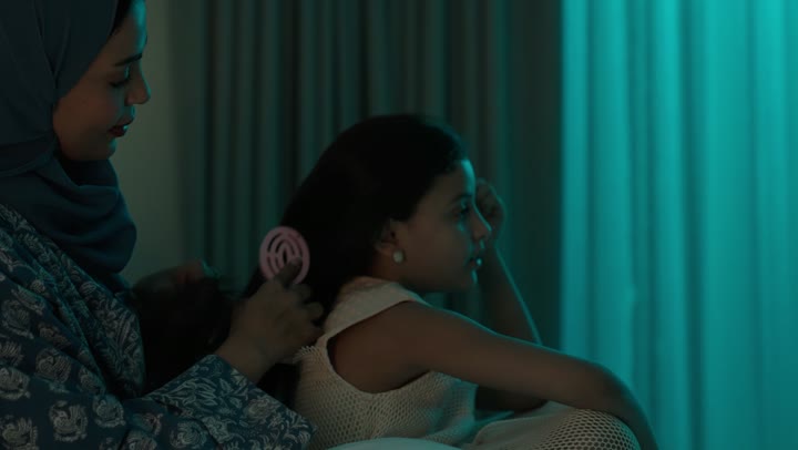 امراة عربية خليجية سعودية ترتدي العباءة والحجاب تجلس على السرير تقوم بتصفيف وتمشيط شعر ابنتها باستخدام الفرشاة ( المشط )، الاهتمام والعناية بالأناقة والمظهر الخارجي، إيماءات السعادة والسرور، المحافظة على شعر صحي وأنيق