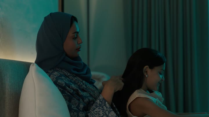 A Saudi Arabian Gulf woman wearing an abaya and hijab sitting on the bed styling and combing her daughter's hair using a brush (comb), taking care and attention to elegance and outward appearance, gestures of happiness and pleasure, maintaining healthy and elegant hair