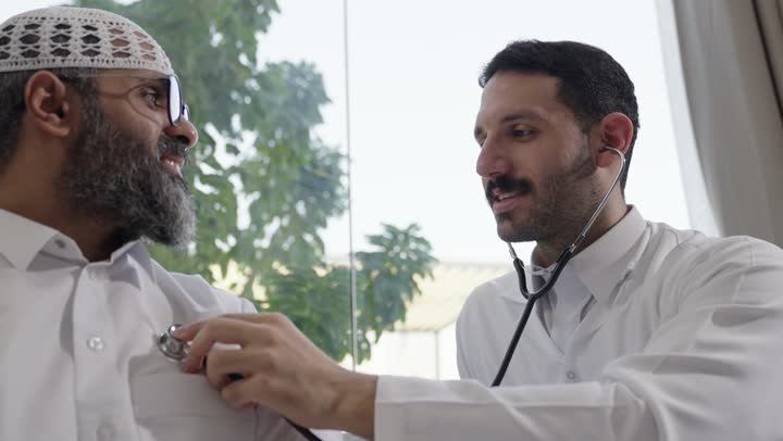 Working in the healthcare sector in Saudi Arabia, providing healthcare and taking care of patients' health, a Saudi Arabian Gulf Arab doctor wearing traditional thobe examining a man with a stethoscope at home, using a stethoscope to hear a heartbeat