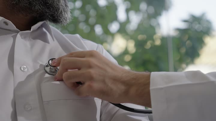 العمل في القطاع الصحي في السعودية،  القيام بالرعاية الصحية والاهتمام بصحة المرضى، طبيب عربي خليجي سعودي يرتدي الثوب التقليدي يقوم بفحص رجل عبر سماعة الأذن في المنزل، استخدام سماعة الطبيب لسماع نبضات القلب