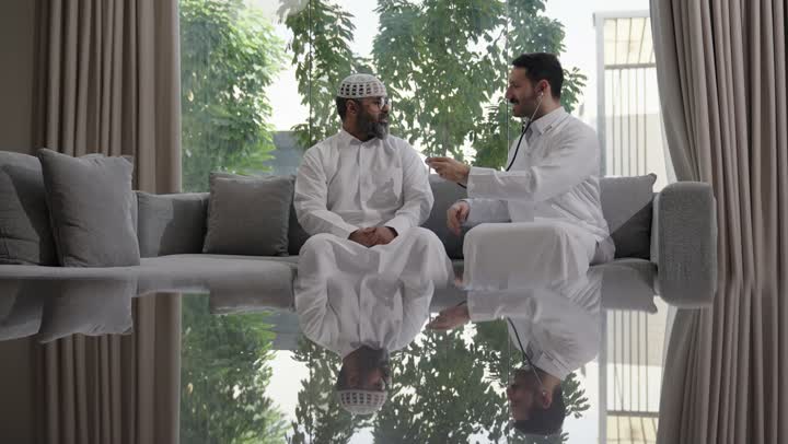 العمل في القطاع الصحي في السعودية،  القيام بالرعاية الصحية والاهتمام بصحة المرضى، طبيب عربي خليجي سعودي يرتدي الثوب التقليدي يقوم بفحص رجل عبر سماعة الأذن في المنزل، استخدام سماعة الطبيب لسماع نبضات القلب