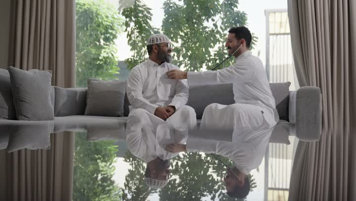 العمل في القطاع الصحي في السعودية،  القيام بالرعاية الصحية والاهتمام بصحة المرضى، طبيب عربي خليجي سعودي يرتدي الثوب التقليدي يقوم بفحص رجل عبر سماعة الأذن في المنزل، استخدام سماعة الطبيب لسماع نبضات القلب