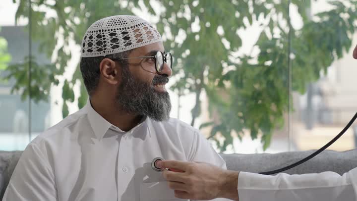 طبيب عربي خليجي سعودي يرتدي الثوب التقليدي يقوم بفحص رجل عبر سماعة الأذن في المنزل، استخدام سماعة الطبيب لسماع نبضات القلب، العمل في القطاع الصحي في السعودية،  القيام بالرعاية الصحية والاهتمام بصحة المرضى