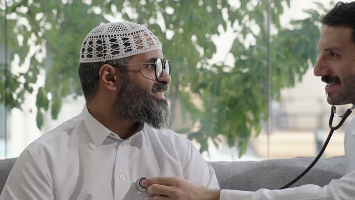 طبيب عربي خليجي سعودي يرتدي الثوب التقليدي يقوم بفحص رجل عبر سماعة الأذن في المنزل، استخدام سماعة الطبيب لسماع نبضات القلب، العمل في القطاع الصحي في السعودية،  القيام بالرعاية الصحية والاهتمام بصحة المرضى