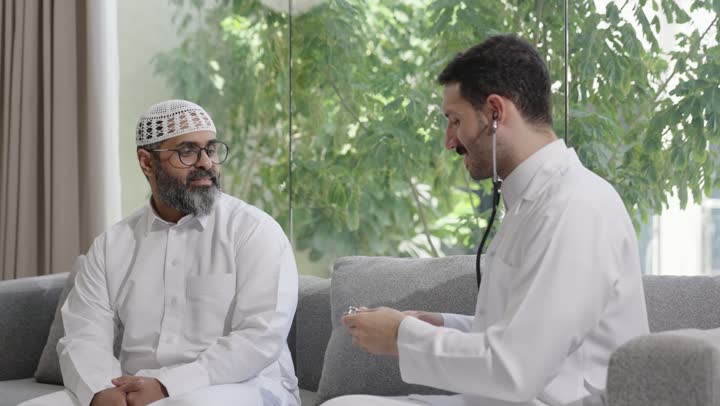 طبيب عربي خليجي سعودي يرتدي الثوب التقليدي يقوم بفحص رجل عبر سماعة الأذن في المنزل، استخدام سماعة الطبيب لسماع نبضات القلب، العمل في القطاع الصحي في السعودية،  القيام بالرعاية الصحية والاهتمام بصحة المرضى