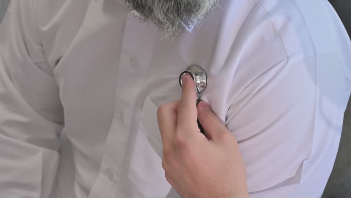 طبيب عربي خليجي سعودي يرتدي الثوب التقليدي يقوم بفحص رجل عبر سماعة الأذن في المنزل، استخدام سماعة الطبيب لسماع نبضات القلب، العمل في القطاع الصحي في السعودية،  القيام بالرعاية الصحية والاهتمام بصحة المرضى