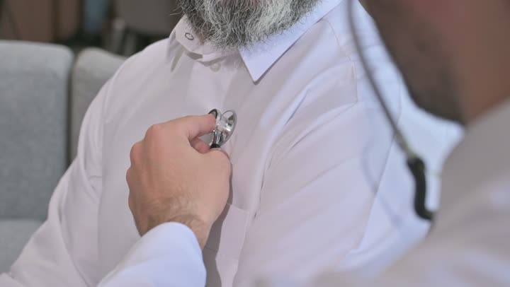 استخدام سماعة الطبيب لسماع نبضات القلب، العمل في القطاع الصحي في السعودية،  القيام بالرعاية الصحية والاهتمام بصحة المرضى، طبيب عربي خليجي سعودي يرتدي الثوب التقليدي يقوم بفحص رجل عبر سماعة الأذن في المنزل