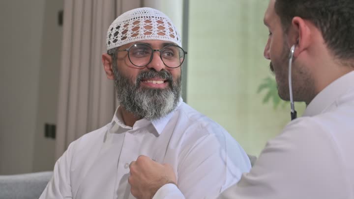استخدام سماعة الطبيب لسماع نبضات القلب، العمل في القطاع الصحي في السعودية،  القيام بالرعاية الصحية والاهتمام بصحة المرضى، طبيب عربي خليجي سعودي يرتدي الثوب التقليدي يقوم بفحص رجل عبر سماعة الأذن في المنزل