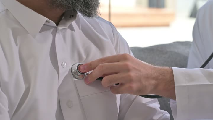 استخدام سماعة الطبيب لسماع نبضات القلب، العمل في القطاع الصحي في السعودية،  القيام بالرعاية الصحية والاهتمام بصحة المرضى، طبيب عربي خليجي سعودي يرتدي الثوب التقليدي يقوم بفحص رجل عبر سماعة الأذن في المنزل