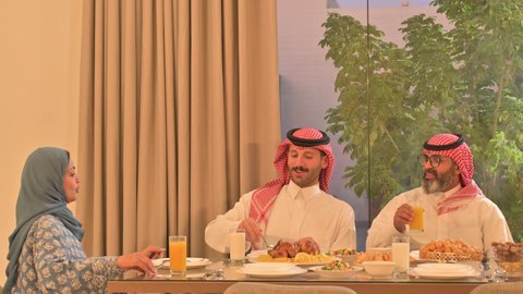 موعد كسر الصيام عند أذان المغرب في رمضان المبارك، فطور رمضاني عائلي، عائلة عربية خليجية سعودية تجلس بايماءا السرور، حسن الضيافة وإكرام الأخرين, أجواء عائلية روحانية دينية إسلامية، اجتماع الأسرة حول مائدة الإفطار المليئة بأصناف المأكولات والمقبلات الشهية