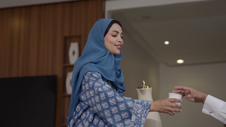 أصالة عادات وتقاليد المجتمع السعودي في المناسبات الاسلامية, كرم الضيافة وحسن الاستقبال في السعودية, شابة عربية خليجية سعودية مبتسمة ترتدي العباءة والحجاب تقوم بصب القهوة الخليجية وتقديمها للضيوف في صالة المنزل, تقديم المشروبات الساخنة العربية الأصيلة في الأعياد