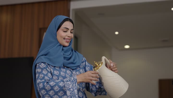 أصالة عادات وتقاليد المجتمع السعودي في المناسبات الاسلامية, كرم الضيافة وحسن الاستقبال في السعودية, شابة عربية خليجية سعودية مبتسمة ترتدي العباءة والحجاب تقوم بصب القهوة الخليجية وتقديمها للضيوف في صالة المنزل, تقديم المشروبات الساخنة العربية الأصيلة في الأعياد
