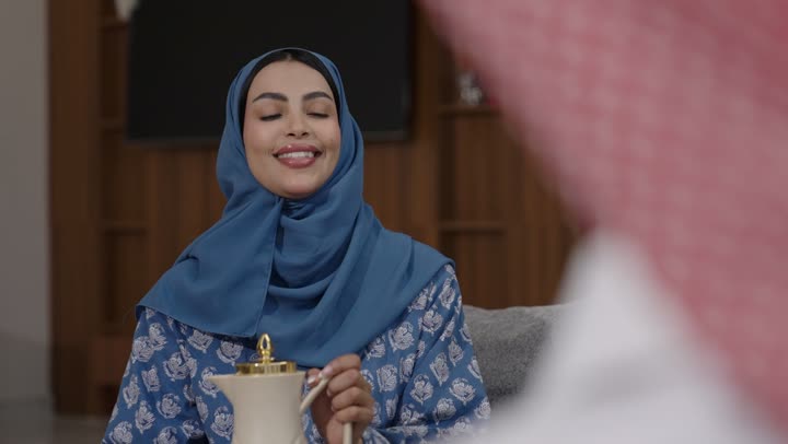 تقديم المشروبات الساخنة العربية الأصيلة في الأعياد، أصالة عادات وتقاليد المجتمع السعودي في المناسبات الاسلامية, كرم الضيافة وحسن الاستقبال في السعودية, شابة عربية خليجية سعودية مبتسمة ترتدي العباءة والحجاب تقوم بصب القهوة الخليجية وتقديمها للضيوف في صالة المنزل