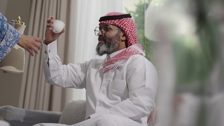 تقديم المشروبات الساخنة العربية الأصيلة في الأعياد، أصالة عادات وتقاليد المجتمع السعودي في المناسبات الاسلامية, كرم الضيافة وحسن الاستقبال في السعودية, شابة عربية خليجية سعودية مبتسمة ترتدي العباءة والحجاب تقوم بصب القهوة الخليجية وتقديمها للضيوف في صالة المنزل