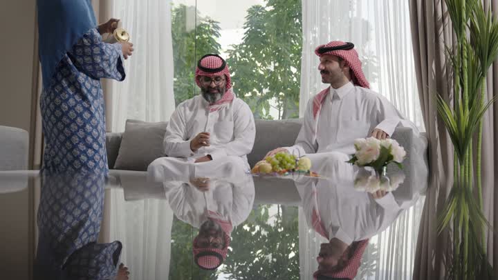 كرم الضيافة وحسن الاستقبال في السعودية, شابة عربية خليجية سعودية مبتسمة ترتدي العباءة والحجاب تقوم بصب القهوة الخليجية وتقديمها للضيوف في صالة المنزل، تقديم المشروبات الساخنة العربية الأصيلة في الأعياد، أصالة عادات وتقاليد المجتمع السعودي في المناسبات الاسلامية