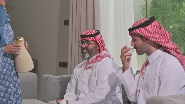 أصالة عادات وتقاليد المجتمع السعودي في المناسبات الاسلامية, كرم الضيافة وحسن الاستقبال في السعودية, شابة عربية خليجية سعودية مبتسمة ترتدي العباءة والحجاب تقوم بصب القهوة الخليجية وتقديمها للضيوف في صالة المنزل, تقديم المشروبات الساخنة العربية الأصيلة في الأعياد