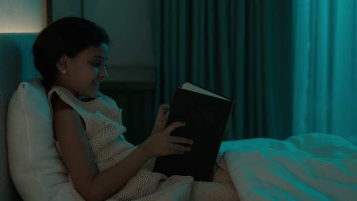 تمضية أوقات الفراغ بالتعلم والتثقف الذاتي, إيماءات الانتباه والتركيز بالمطالعة, تصوير مقرب لفتاة عربية خليجية سعودية تجلس على السرير تقوم بالقراءة بتمعن قبل النوم , مطالعة القصص التصويرية المسلية, تنمية مهارات القراءة للأطفال, الاهتمام بتصفح الكتب المفيدة