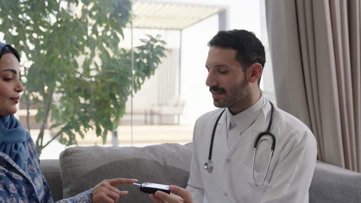 شابة سعودية مصابة بحالة مرضية مزمنة, المتابعة الدورية لمرضى داء السكري, الفحوصات الطبية السريعة لمعرفة نسبة السكر في الدم، استخدام الأجهزة الالكترونية للتحاليل المنزلية، تصوير مقرب لطبيب عربي خليجي سعودي يرتدي المعطف الابيض وسماعة الأذن يقوم بوخز اصبع امرأة