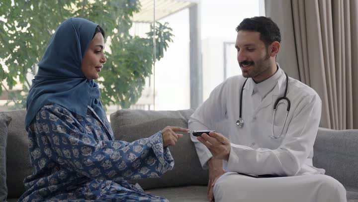شابة سعودية مصابة بحالة مرضية مزمنة, المتابعة الدورية لمرضى داء السكري, الفحوصات الطبية السريعة لمعرفة نسبة السكر في الدم، استخدام الأجهزة الالكترونية للتحاليل المنزلية، تصوير مقرب لطبيب عربي خليجي سعودي يرتدي المعطف الابيض وسماعة الأذن يقوم بوخز اصبع امرأة