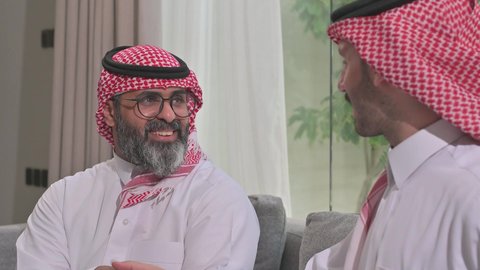 Chatting and exchanging conversations, a happy family gathering, expressions of affection and love, enjoying spending time with the children, an elderly Saudi Arabian Gulf man wearing the traditional thobe and shemagh sitting on the sofa next to his young son in the living room