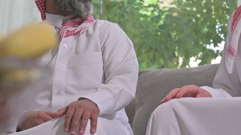 التسامر وتبادل الاحاديث، جلسة عائلية سعيدة، تعابير المودة والمحبة، الاستمتاع بقضاء وقت مع الابناء، رجل كبير بالسن عربي خليجي سعودي يرتدي الثوب التقليدي والشماغ يجلس على الاريكة بجانب ابنه الشاب في غرفة الجلوس
