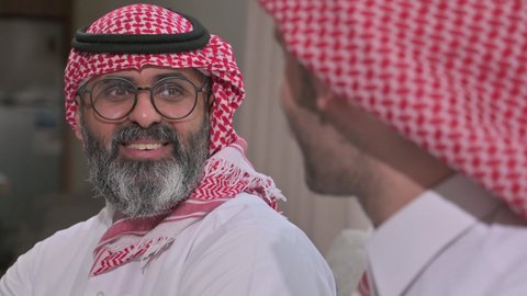  الاستمتاع بقضاء وقت مع الابناء، رجل كبير بالسن عربي خليجي سعودي يرتدي الثوب التقليدي والشماغ يجلس على الاريكة بجانب ابنه الشاب في غرفة الجلوس، التسامر وتبادل الاحاديث، جلسة عائلية سعيدة، تعابير المودة والمحبة