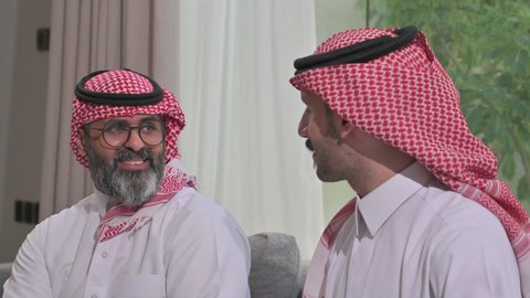 التسامر وتبادل الاحاديث، جلسة عائلية سعيدة، تعابير المودة والمحبة، الاستمتاع بقضاء وقت مع الابناء، رجل كبير بالسن عربي خليجي سعودي يرتدي الثوب التقليدي والشماغ يجلس على الاريكة بجانب ابنه الشاب في غرفة الجلوس