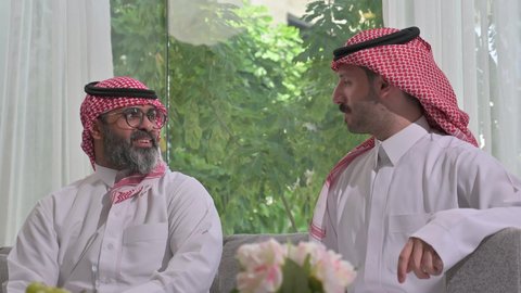  الاستمتاع بقضاء وقت مع الابناء، رجل كبير بالسن عربي خليجي سعودي يرتدي الثوب التقليدي والشماغ يجلس على الاريكة بجانب ابنه الشاب في غرفة الجلوس، التسامر وتبادل الاحاديث، جلسة عائلية سعيدة، تعابير المودة والمحبة