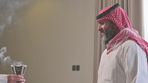 عادات وتقاليد المجتمع السعودي في الأعياد والمناسبات الاسلامية، الاصالة والعراقة والتراث السعودي, عطورات وروائح خليجية مميزة، تصوير من الجانب لرجل شايب كبير بالسن عربي خليجي سعودي يرتدي الثوب التقليدي والشماغ يقوم بتبخير نفسه بالمبخرة، التبخر والتطيب بخشب العود