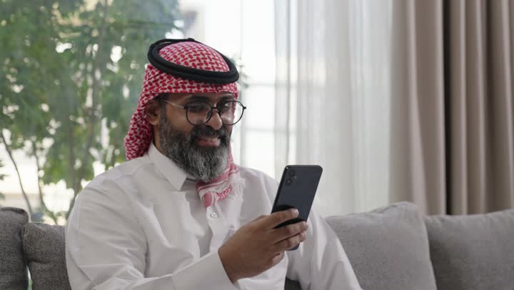 مفهوم الحب والحنان، استخدام الهاتف المحمول، تطور التكنولوجيا والأجهزة التقنية، التسوق الالكتروني وتصفح برامج وتطبيقات الجوال، تصوير مقرب لرجل عربي خليجي سعودي يرتدي الثوب التقليدي والشماغ يجلس في صالون المنزل يقوم باحتضان ابنته ، الدردشة مع الأهل والأصدقاء