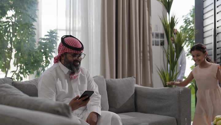 مفهوم الحب والحنان، استخدام الهاتف المحمول، تطور التكنولوجيا والأجهزة التقنية، التسوق الالكتروني وتصفح برامج وتطبيقات الجوال، تصوير مقرب لرجل عربي خليجي سعودي يرتدي الثوب التقليدي والشماغ يجلس في صالون المنزل يقوم باحتضان ابنته ، الدردشة مع الأهل والأصدقاء